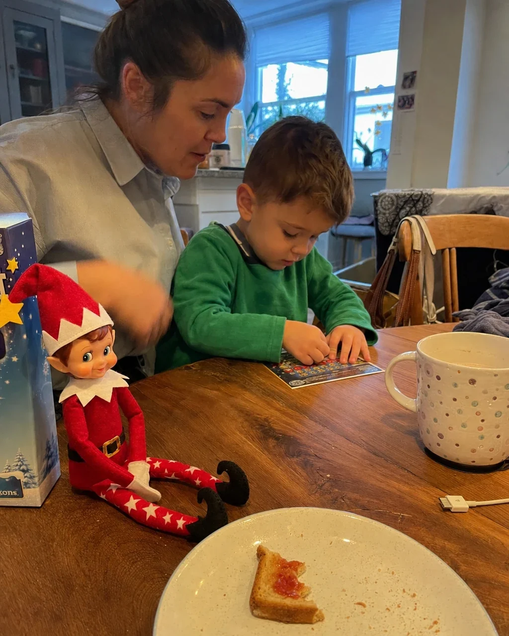 A mother in her work shirt beside her son at the breakfast table as he scratches a card