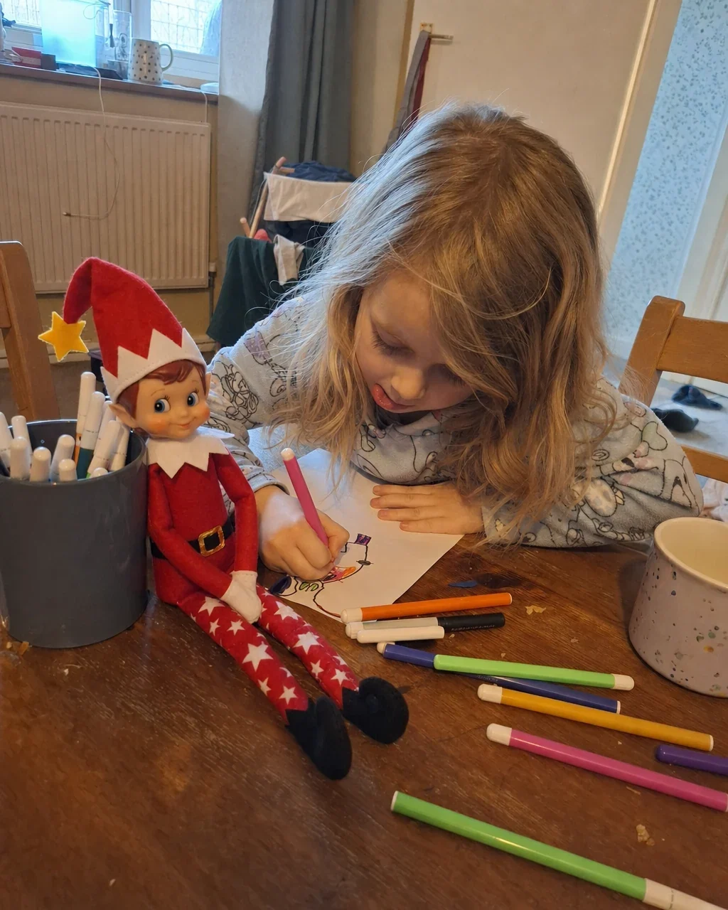 A child drawing at the dining table with felt-tip pens, Pip propped against the pen pot